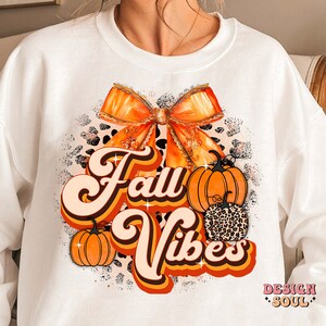 May include: White sweatshirt with a graphic design featuring an orange bow, pumpkins, and the text "Fall Vibes" in a retro font.
