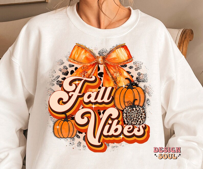 May include: White sweatshirt with a graphic design featuring an orange bow, pumpkins, and the text "Fall Vibes" in a retro font.