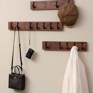 Könnte beinhalten: Drei Wand-Kleiderhaken aus Holz mit Haken. Der obere Haken hat zwei Haken, der mittlere Haken hat acht Haken und der untere Haken hat vier Haken. Ein brauner Hut hängt am oberen Haken, eine schwarze Handtasche hängt am mittleren Haken, eine Kamera hängt am mittleren Haken und ein weißer Schal hängt am unteren Haken.