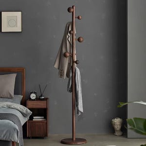 Op de afbeelding: Een donkerbruine houten kapstok met meerdere afgeronde knoppen om items op te hangen. Een beige blazer en een grijze sjaal hangen aan de kapstok. De kapstok staat op een ronde basis. De achtergrond is een donkergrijze muur.