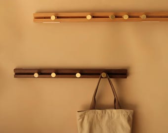 May include: Two wooden coat racks with brass knobs are mounted on a beige wall. The top rack is light-colored wood, and the bottom rack is dark wood. A canvas tote bag hangs from one of the knobs on the bottom rack.