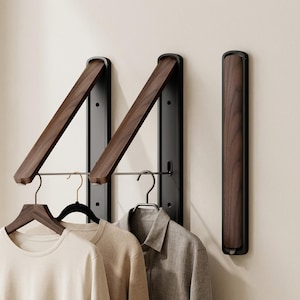 Puede incluir: Tres percheros de pared con base de metal negro y brazo de madera marrón oscuro con veteado. Dos percheros están extendidos hacia afuera con una barra metálica horizontal que sostiene perchas, mientras que uno está plegado contra la pared.
