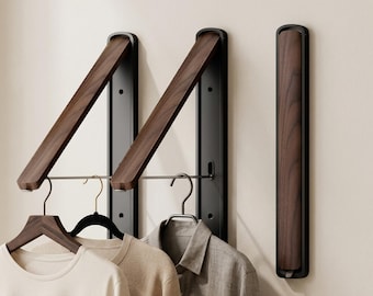 May include: Wall-mounted folding clothes racks featuring a dark wood grain finish and black metal frames. Two racks are extended outward with metal rods holding wooden and black velvet hangers with shirts, while one rack is folded flat against the wall.