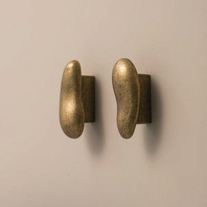 Puede incluir: Dos ganchos de pared con acabado en latón. Cada gancho presenta una forma orgánica y lisa similar a un guijarro que se extiende desde una base rectangular. La superficie metálica tiene una textura mate y ligeramente desgastada en tonos dorados.