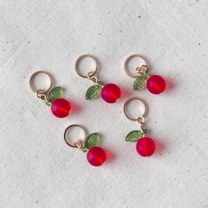 May include: Five gold-toned stitch markers, each featuring a red bead cherry with green leaf accents. These small, decorative charms are designed for use in knitting or crochet projects, adding a touch of whimsy to crafting.