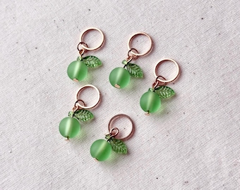 May include: Five green apple-shaped stitch markers with copper-colored rings and leaf accents. The markers are made of frosted glass and are arranged on a white surface. These markers are ideal for knitting and crochet projects.