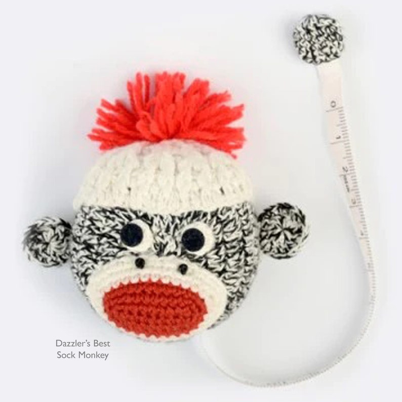 May include: A handmade sock monkey tape measure with a white and black body, red mouth, and orange yarn hair. The tape measure extends from the monkey's head. The text "Dazzler's Best Sock Monkey" is visible.