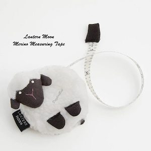 May include: A white sheep-shaped measuring tape with black accents and a black tag that reads "Lantern Moon". The measuring tape extends from the sheep's body, showing inch measurements. The text "Merino Measuring Tape" is also visible.