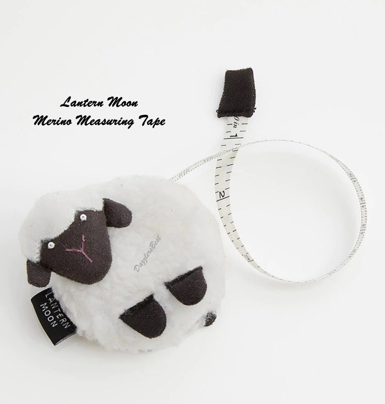 May include: A white sheep-shaped measuring tape with black accents and a black tag that reads "Lantern Moon". The measuring tape extends from the sheep's body, showing inch measurements. The text "Merino Measuring Tape" is also visible.