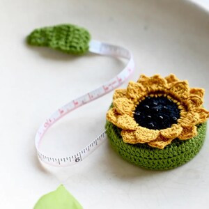 May include: A crocheted sunflower tape measure with a green base and yellow petals. The tape extends from a green leaf, with measurements in inches. The tape measure is a unique and decorative tool for sewing and crafting projects.
