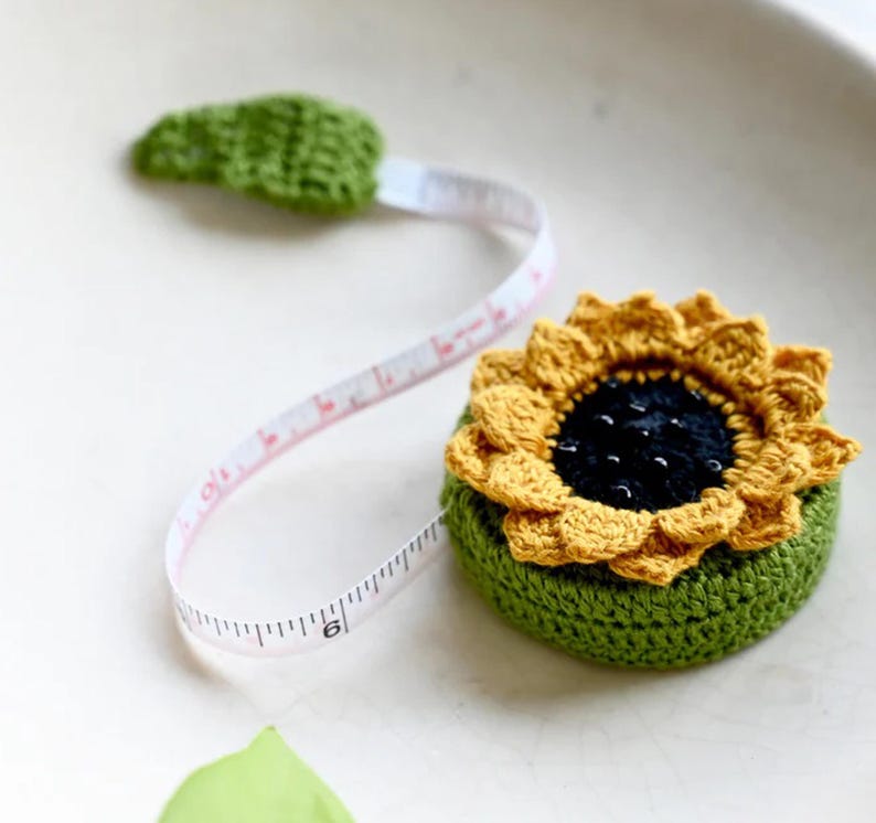 May include: A crocheted sunflower tape measure with a green base and yellow petals. The tape extends from a green leaf, with measurements in inches. The tape measure is a unique and decorative tool for sewing and crafting projects.