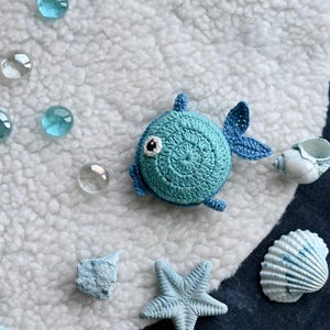 May include: A crocheted blue fish with white and black eye details, fins, and tail. The fish is surrounded by glass beads, seashells, and a starfish on a white and blue striped surface. The fish is a handmade item.