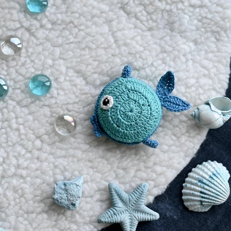 May include: A crocheted blue fish with white and black eye details, fins, and tail. The fish is surrounded by glass beads, seashells, and a starfish on a white and blue striped surface. The fish is a handmade item.