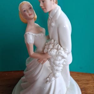May include: A white porcelain wedding cake topper featuring a bride and groom in an embrace. The bride wears a white gown and holds a bouquet. The groom is in a white suit. The base is round and decorated with embossed details.