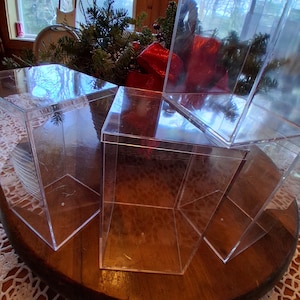 May include: A collection of clear, cube-shaped display boxes are positioned on a circular wooden surface. The transparent boxes offer a clear view of their contents. The background features a window and festive decorations.