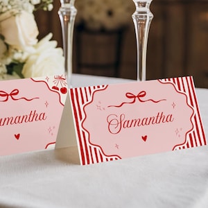 Puede incluir: Dos tarjetas de mesa rosas y rojas con el nombre "Samantha" en escritura elegante. Una tarjeta presenta un borde decorativo con corazones y lazos, mientras que la otra tiene rayas rojas. Las tarjetas están sobre un mantel blanco, con flores y cristalería de cristal en el fondo.