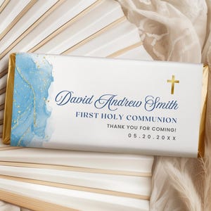May include: A chocolate bar wrapper with a blue and gold watercolour design. The wrapper features the text "David Andrew Smith", "First Holy Communion", and a gold cross. The date 05.20.20XX is also printed on the wrapper.