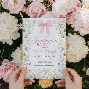 May include: A graduation party invitation with a pink bow and floral border. The invitation reads "Join us for a Graduation Party in Honor of Sarah Michelle." The date is Saturday, June 13th, at 10:00 am. The invitation is held in front of a backdrop of flowers.