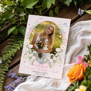 May include: A graduation announcement card with a photograph of a young woman holding flowers, surrounded by a floral border. The card displays the name "Sarah Michelle Smith", school details, and "Class of 2026", with a pink bow at the bottom.