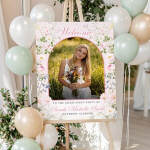 May include: A graduation welcome sign on an easel. The sign has a photo of a young woman with flowers, floral designs, and the word "Welcome". White, green, and tan balloons are arranged nearby. The sign is for Sarah Michelle Smith.