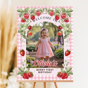 Op de afbeelding: Een roze en witte geruite welkomstbord met een foto van een lachend kind in een roze jurk. Het bord is versierd met aardbeienillustraties en de tekst "Welcome to Olivia's Berry First Birthday."
