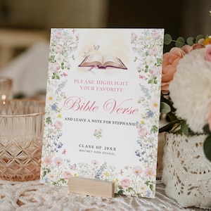 May include: A floral-themed Bible Verse sign with the text "PLEASE HIGHLIGHT YOUR FAVORITE Bible Verse AND LEAVE A NOTE FOR STEPHANIE" and "CLASS OF 20XX WHITNEY HIGH SCHOOL". The sign is propped up by a small wooden block.