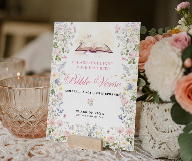 May include: A floral-themed Bible Verse sign with the text "PLEASE HIGHLIGHT YOUR FAVORITE Bible Verse AND LEAVE A NOTE FOR STEPHANIE" and "CLASS OF 20XX WHITNEY HIGH SCHOOL". The sign is propped up by a small wooden block.