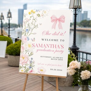 May include: A white graduation party welcome sign on a wooden easel. The sign features a pink bow and floral decorations. Text reads "She did it! Welcome to Samantha's graduation party" with the date and school name.