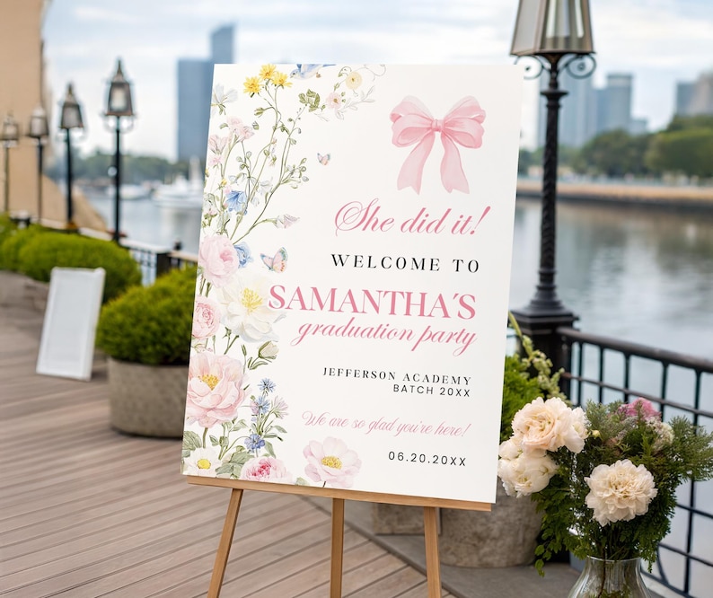 May include: A white graduation party welcome sign on a wooden easel. The sign features a pink bow and floral decorations. Text reads "She did it! Welcome to Samantha's graduation party" with the date and school name.