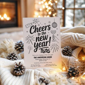 May include: A white New Year's invitation card with black text and illustrations. The card reads "Cheers to the new year! 2026" with details for a party at The Anderson Home. The card is surrounded by pinecones and a cozy blanket.