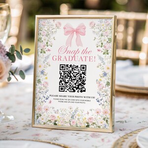 May include: A gold-framed sign with the text "Snap the GRADUATE!" and a QR code, decorated with flowers and a pink bow. The sign is on a table with a floral tablecloth. The sign encourages photo sharing.