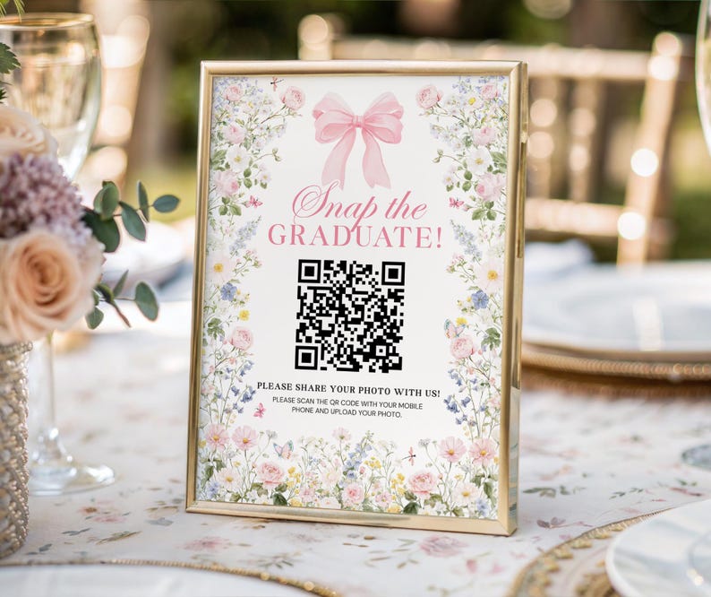 May include: A gold-framed sign with the text "Snap the GRADUATE!" and a QR code, decorated with flowers and a pink bow. The sign is on a table with a floral tablecloth. The sign encourages photo sharing.