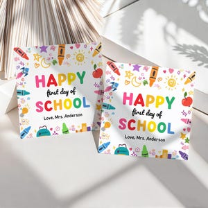 May include: Two square cards with the text "HAPPY first day of SCHOOL" in colorful letters. The cards are decorated with illustrations of crayons, apples, rockets, and stars. The text "Love, Mrs. Anderson" is also included.