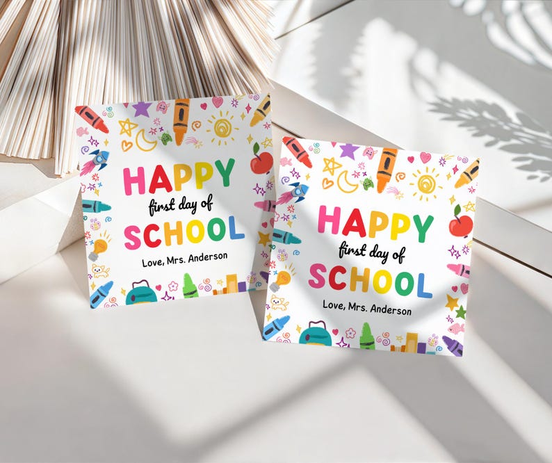 May include: Two square cards with the text "HAPPY first day of SCHOOL" in colorful letters. The cards are decorated with illustrations of crayons, apples, rockets, and stars. The text "Love, Mrs. Anderson" is also included.