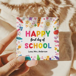 May include: A square card with the words "HAPPY first day of SCHOOL" in colorful letters, surrounded by illustrations of crayons, stars, and an apple. The text "Love, Mrs. Anderson" is also included.