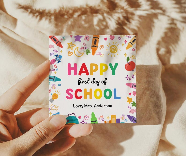 May include: A square card with the words "HAPPY first day of SCHOOL" in colorful letters, surrounded by illustrations of crayons, stars, and an apple. The text "Love, Mrs. Anderson" is also included.