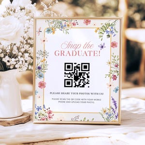 May include: A framed sign with floral accents and the text "Snap the GRADUATE!". The sign includes a QR code and instructions to share photos. The frame is gold, and the flowers are in shades of pink, blue, and purple.