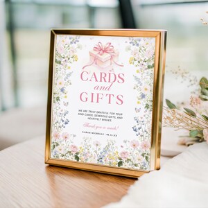 May include: A gold-framed sign with the text "CARDS and GIFTS" in pink. The sign features a floral border and a gift box illustration. The text reads "We are truly grateful for your kind cards, generous gifts, and heartfelt wishes."