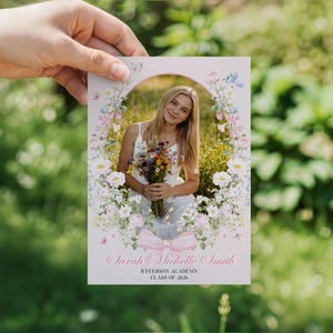 May include: A graduation announcement card with a floral design and a photo of a young woman holding flowers. The text reads "Sarah Michelle Smith, Jefferson Academy, Class of 2026." The card is held against a blurred green background.