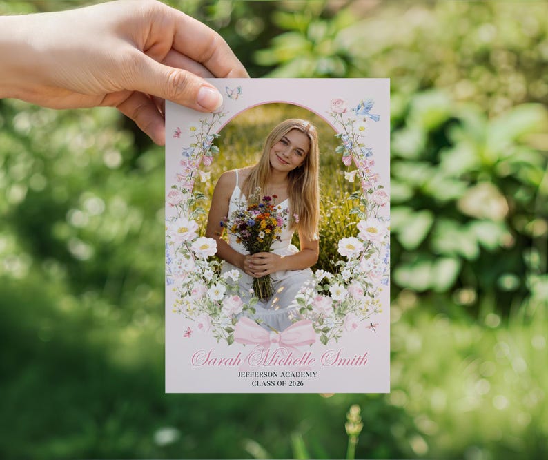 May include: A graduation announcement card with a floral design and a photo of a young woman holding flowers. The text reads "Sarah Michelle Smith, Jefferson Academy, Class of 2026." The card is held against a blurred green background.