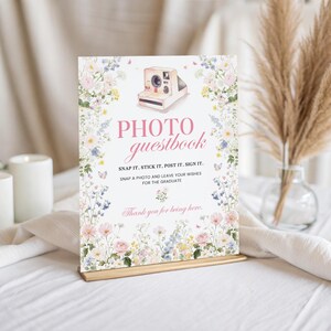 May include: A rectangular photo guestbook sign with floral and butterfly illustrations. The sign reads "PHOTO guestbook" and includes instructions to snap, stick, post, and sign. The sign is on a gold stand, with candles and pampas grass in the background.