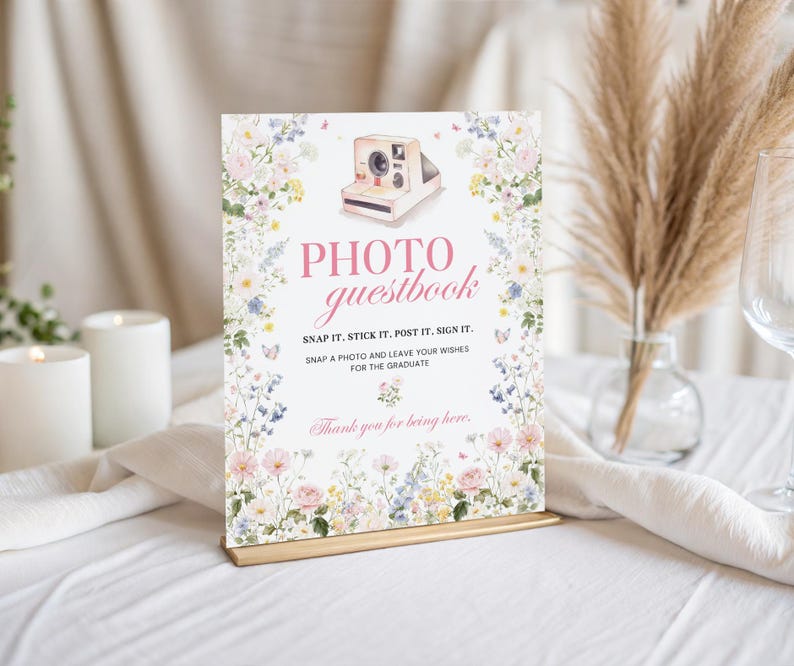 May include: A rectangular photo guestbook sign with floral and butterfly illustrations. The sign reads "PHOTO guestbook" and includes instructions to snap, stick, post, and sign. The sign is on a gold stand, with candles and pampas grass in the background.