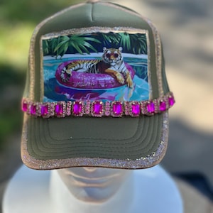 May include: An olive green trucker cap featuring a rectangular graphic of a tiger wearing sunglasses on a pink glittery pool float. The cap is accented with a row of rectangular hot pink rhinestones and a shimmering glitter trim along the brim.