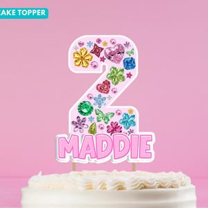 Peut inclure: Une décoration de gâteau en forme de chiffre deux, avec un fond rose clair orné de diverses fausses pierres précieuses colorées en forme de fleurs, de cœurs et de papillons. Le prénom MADDIE est inscrit en lettres roses épaisses.