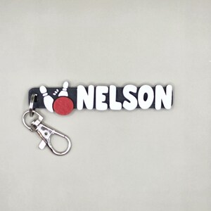 Puede incluir: Llavero negro y blanco con el nombre "NELSON" en letras blancas y un gráfico de bola de bolos roja. El llavero tiene un cierre de metal plateado.