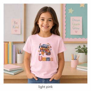 Puede incluir: Una camiseta de manga corta de color rosa claro con un gráfico de libros apilados, un lápiz, tijeras y una mochila. Debajo del gráfico, el nombre Umma está impreso en letras grandes, coloridas y estampadas. La camiseta tiene cuello redondo.