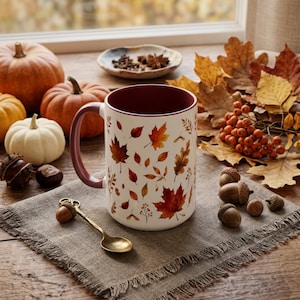 May include: A white ceramic mug with a maroon interior and handle, decorated with a pattern of colourful autumn leaves. The mug sits on a grey linen napkin next to a small gold-toned spoon, surrounded by pumpkins, acorns, and dried leaves.