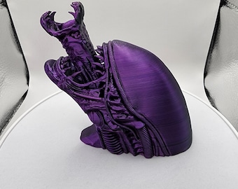 May include: A purple, 3D-printed sculpture of an alien head, featuring intricate details and a smooth, curved back. The sculpture has a textured, open-mouthed design with visible ribbing and a pointed head.