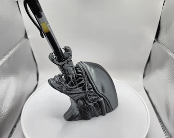 May include: A black 3D printed pen holder shaped like an alien head with a pen sticking out of its mouth.