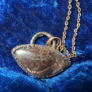 May include: A copper-toned pendant necklace with an oval, textured centerpiece. The pendant has a detailed border and a chain with a matching copper finish. The background is a deep blue velvet.